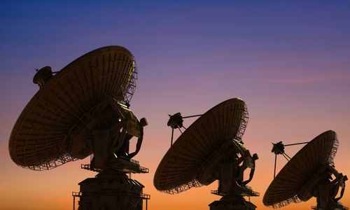 Three large satellite dishes are silhouetted against a vibrant sunset sky