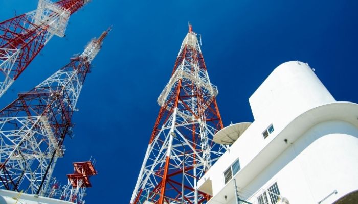 Three towering red and white communication towers rise against a clear blue sky beside a white building.