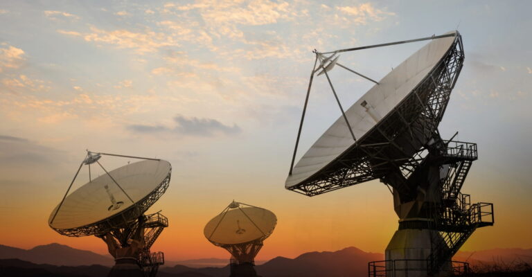 Three large satellite dishes silhouetted against a vibrant sunset in a desert landscape